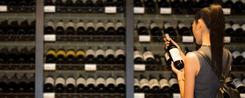 header image: A woman with black hair in a wine shop holding a wine bottle and reading the label